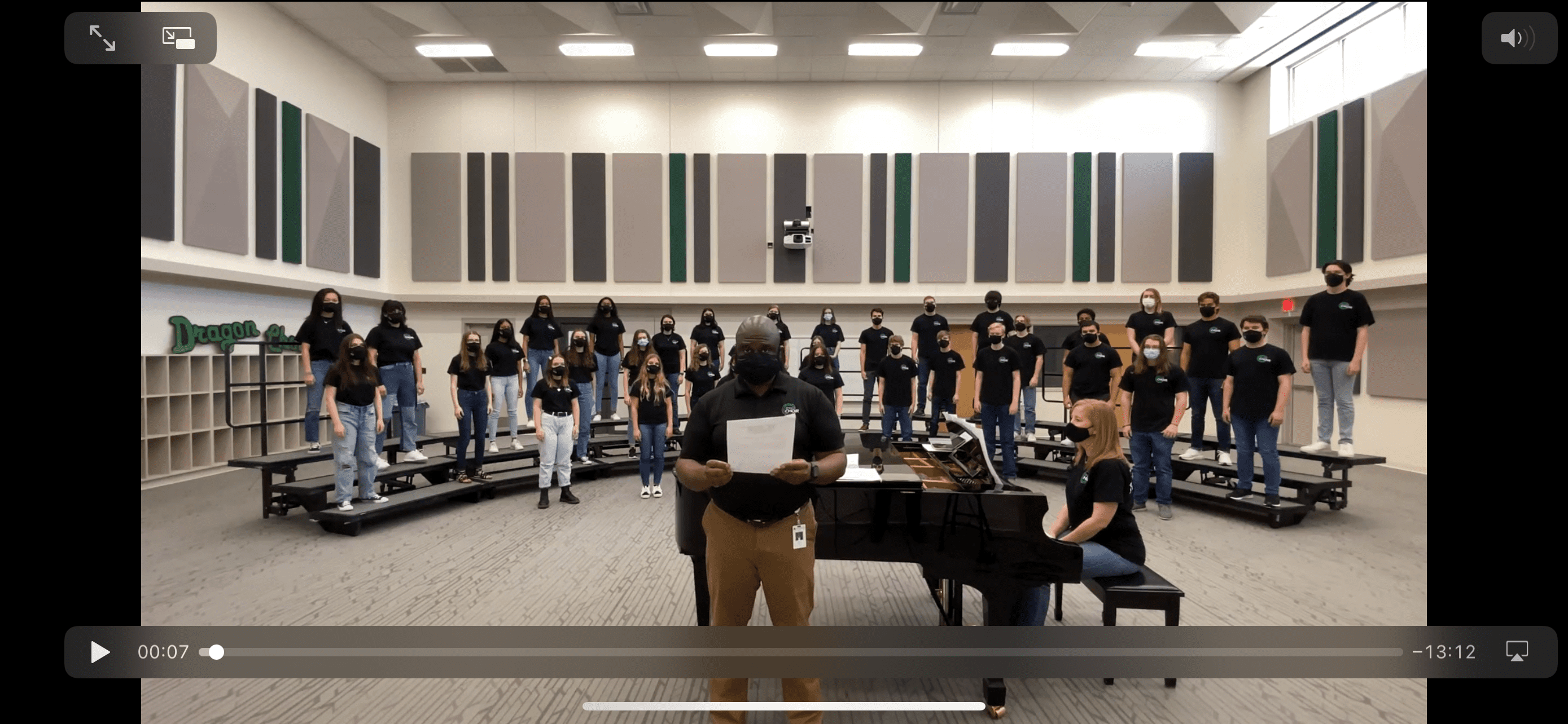 A choir is performing in a brightly lit room. The conductor stands in front holding papers, while a pianist sits at a black grand piano. The choir members, wearing black shirts and jeans, are standing on tiered risers in the background.