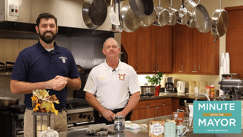 Two men stand in a kitchen, smiling. One wears a navy shirt with a logo, while the other wears a white shirt with a fire department badge. Pans hang above, and a coffee maker sits on the counter. A "Minute with the Mayor" graphic is in the corner.