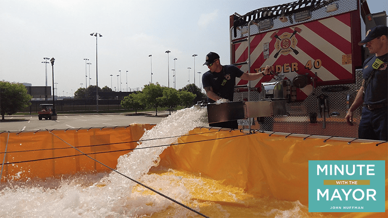Firefighters in uniforms stand by a fire truck marked "Tender 40," guiding water into a large yellow reservoir. The scene takes place in a parking lot. A graphic at the bottom right reads, "Minute with the Mayor John Huffman.