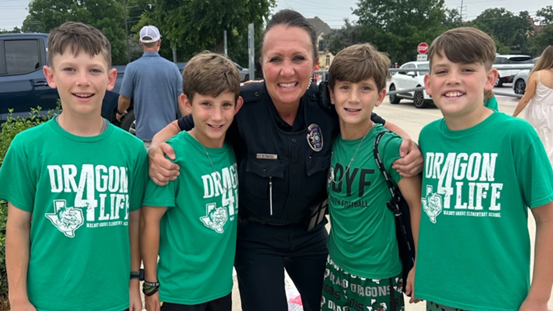A police officer stands smiling with four boys, all wearing green "Dragon for Life" shirts. They are outside, with trees and parked cars visible in the background. The group appears happy and is posing for the camera.