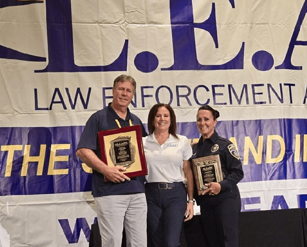 Three people stand in front of a large banner that reads "L.E.A.P. Law Enforcement." The two individuals on the left hold plaques, while the person on the right is in a police uniform. They are posing and smiling for the photo.