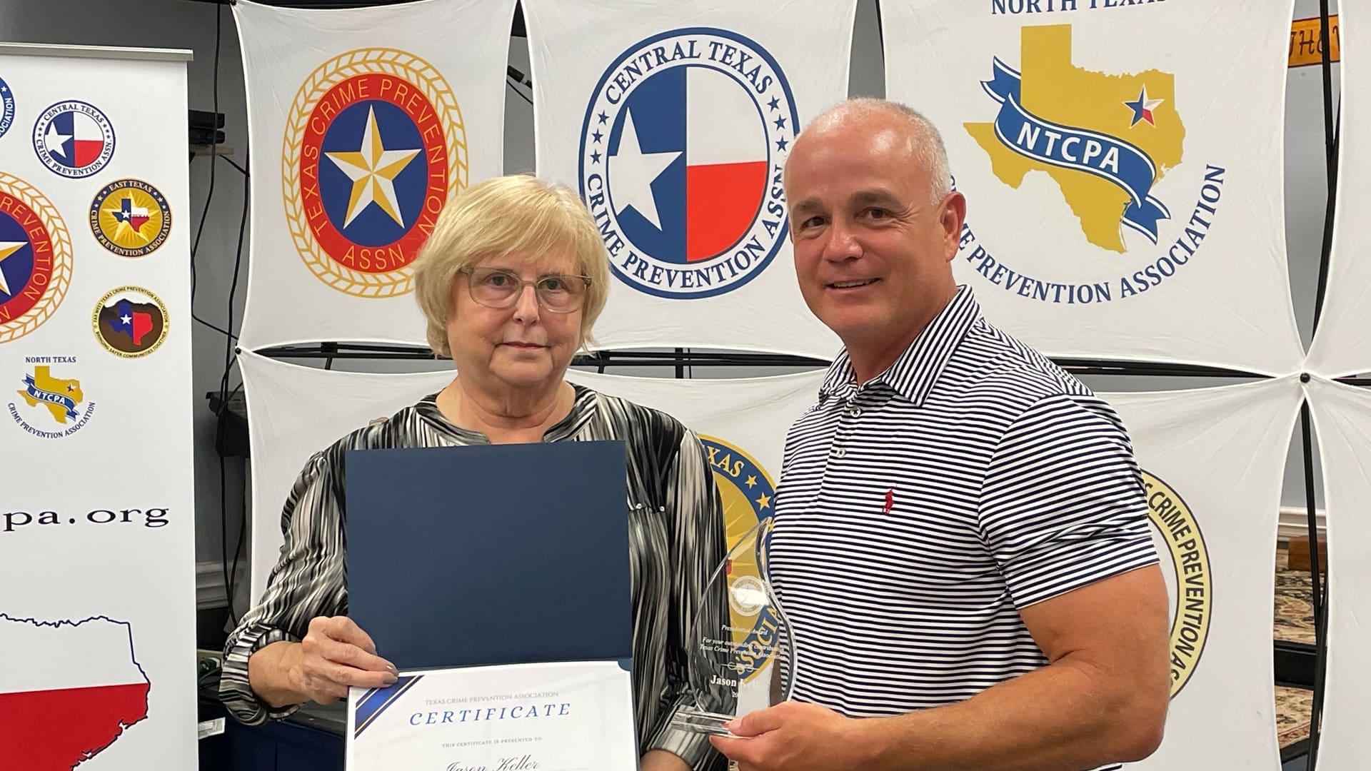 Two people stand indoors. The older person on the left holds a certificate, while the person on the right holds a small plaque. Behind them are banners featuring various Texas and crime prevention logos. They both look towards the camera.