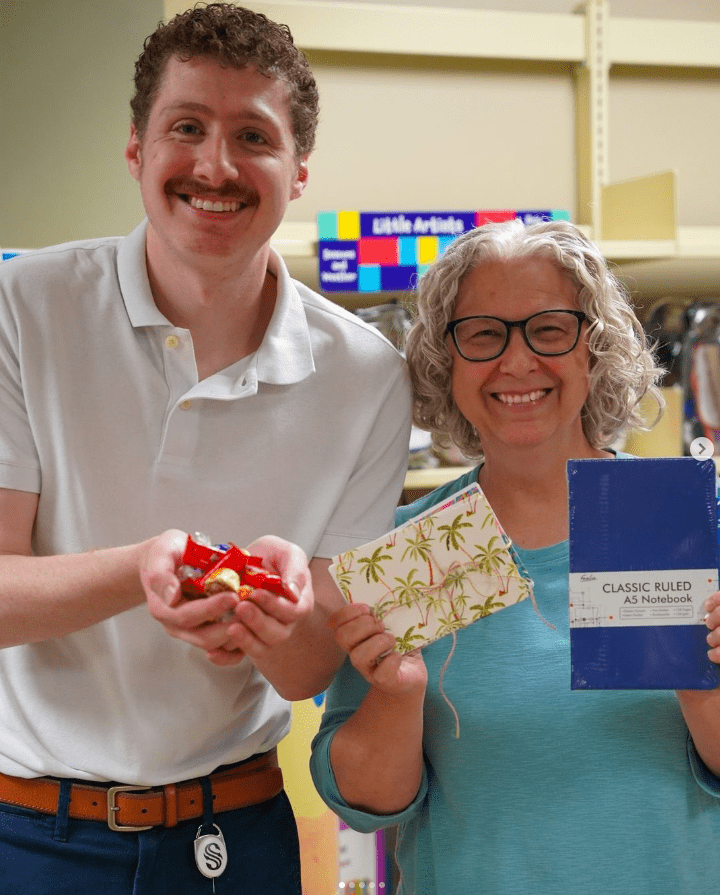 A smiling man and woman pose together indoors. The man holds a handful of small red-wrapped items. The woman holds a palm tree patterned pouch and a blue notebook. Shelves with colorful items are in the background.