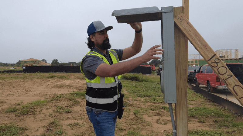 A man in a safety vest and blue cap inspects or installs an electrical panel on a wooden post at a construction site with vehicles and partially built houses in the background.