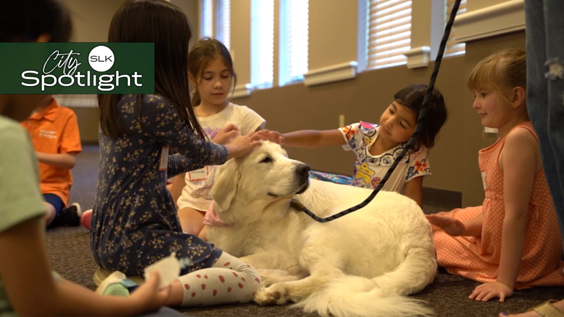 A group of young children sit on the floor, petting a calm, white dog lying down indoors. Sunlight streams through nearby windows. A graphic in the upper left corner reads "City Spotlight SLK.