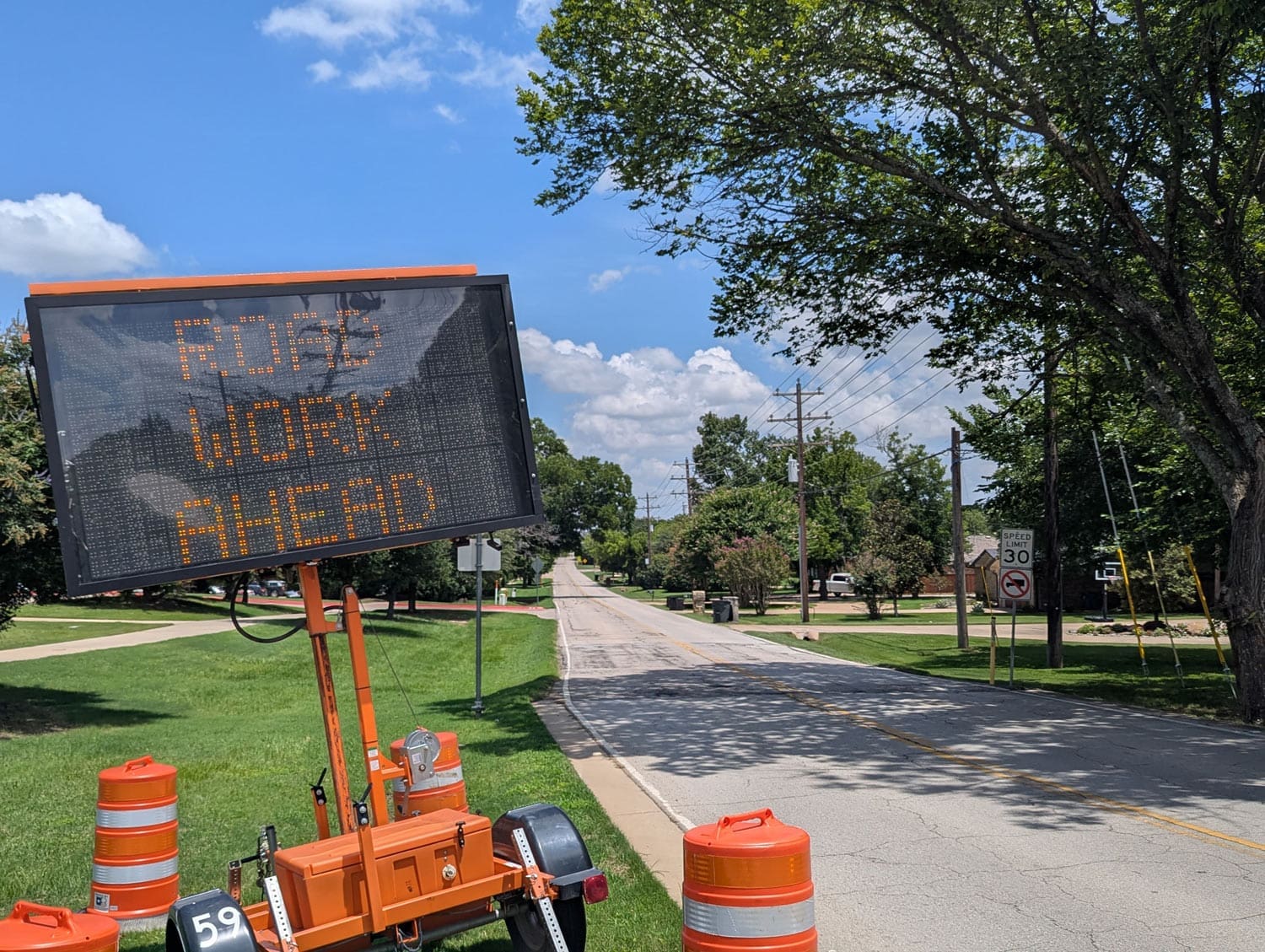 An electronic sign beside a road displays "ROAD WORK AHEAD." Orange traffic barrels line the roadside, and trees, grass, and houses are visible along the street under a blue sky with clouds.