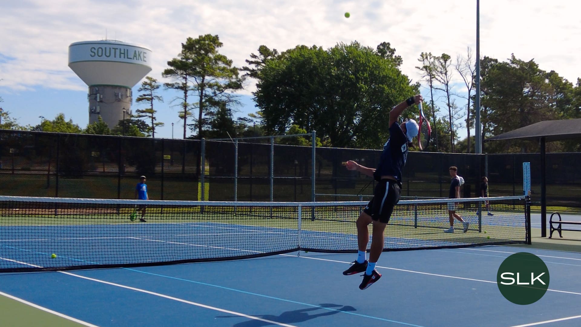 A tennis player jumps to hit a serve on an outdoor court. Other players and a coach stand nearby. Trees and a large water tower labeled "SOUTHLAKE" are visible in the background. The SLK logo appears in the corner.