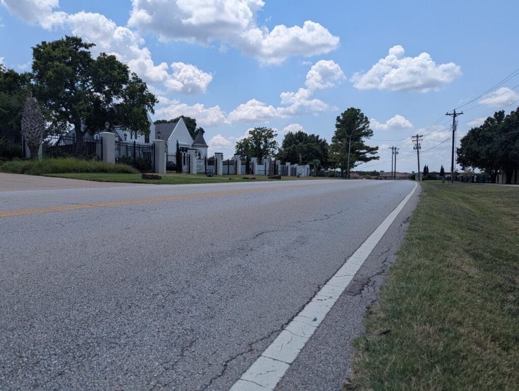 A quiet, empty road curves slightly, bordered by grass and trees on a sunny day with a blue sky and scattered clouds. A row of white buildings with fences is visible on the left side of the road.