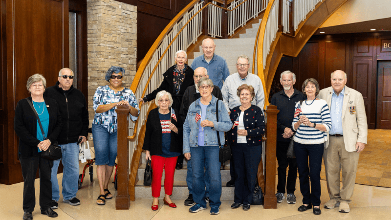 A group of older adults pose and smile together in front of a staircase inside a building. Most are standing, some hold small American flags, and everyone is dressed in casual attire.