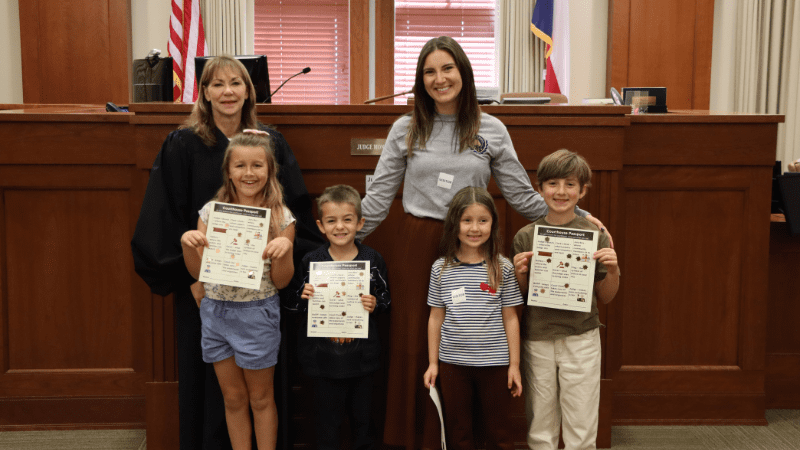 A judge and an adult stand with four smiling children holding certificates in a courtroom. The judge wears a black robe, and flags are visible in the background near the judge's bench.