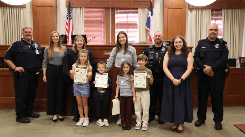 A group of children holding certificates stands with several adults, including police officers in uniform, in a courtroom setting with American and Texas flags in the background.