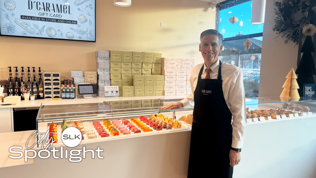 Mayor Shawn McCaskill in a black apron stands behind a pastry display case filled with colorful desserts in a modern bakery. Gift boxes are stacked behind him, and a sign advertises D'Caramel gift cards. The "SLK Spotlight" logo is visible.