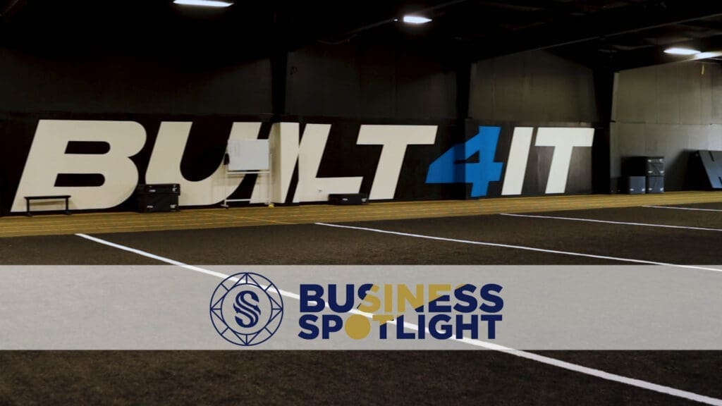 Indoor sports facility with large "BUILT 4 IT" mural on the wall, artificial turf field, and a "Business Spotlight" banner with a circular logo in the foreground.