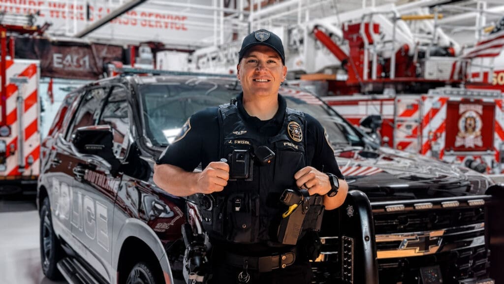 A police officer in uniform stands smiling in front of a black patrol SUV inside a brightly lit fire station with fire trucks in the background.