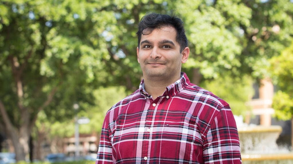 A person wearing a red plaid shirt stands outdoors in front of green trees, smiling slightly at the camera. The background is blurred with sunlight filtering through the leaves.