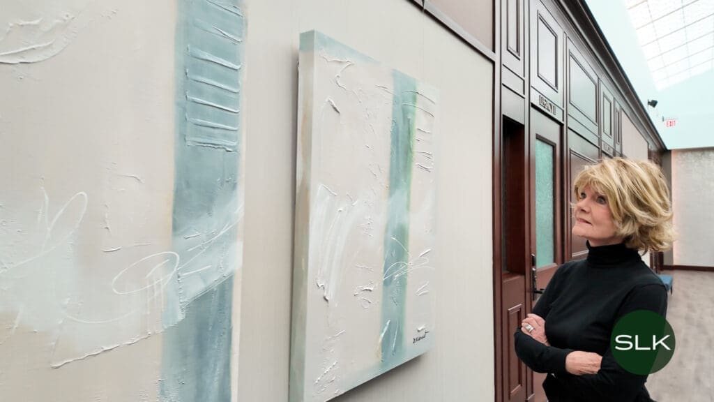 A woman with short blonde hair and a black turtleneck stands in a hallway, thoughtfully observing two abstract paintings with blue and white brushstrokes. Doors and a green SLK logo are visible in the background.
