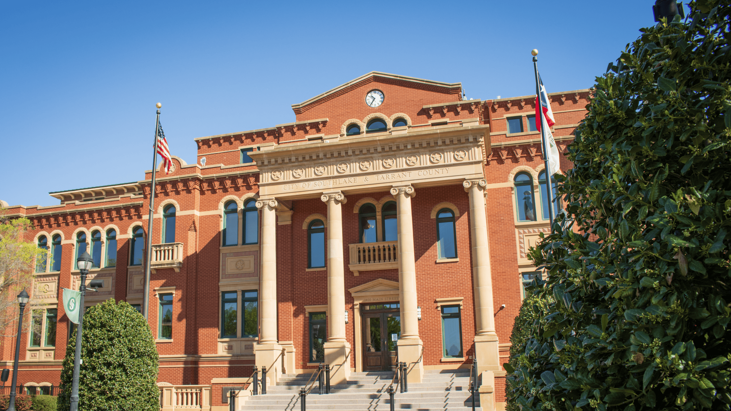 Historic red-brick Southlake Town Hall building with tall stone columns, arched windows, and a clock centered above the main entrance. American, Texas, and city flags flank the building, with manicured trees and shrubs framing the front under a clear blue sky.