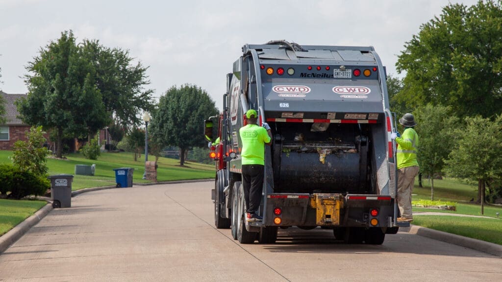 Two sanitation workers in green shirts stand on the back of a garbage truck as it moves through a suburban neighborhood, collecting trash bins on a sunny day.